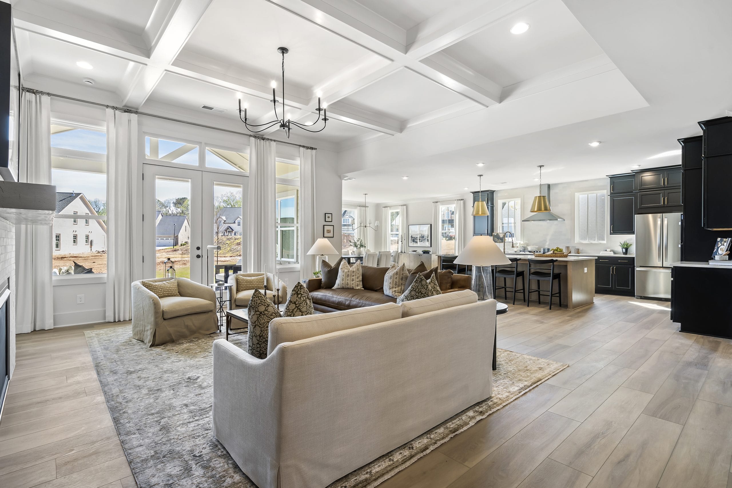 Spacious open-concept living area with living room, dining area, and kitchen, featuring large rug, sofas, throw pillows, chandelier, modern appliances, kitchen island with bar stools, and large windows.