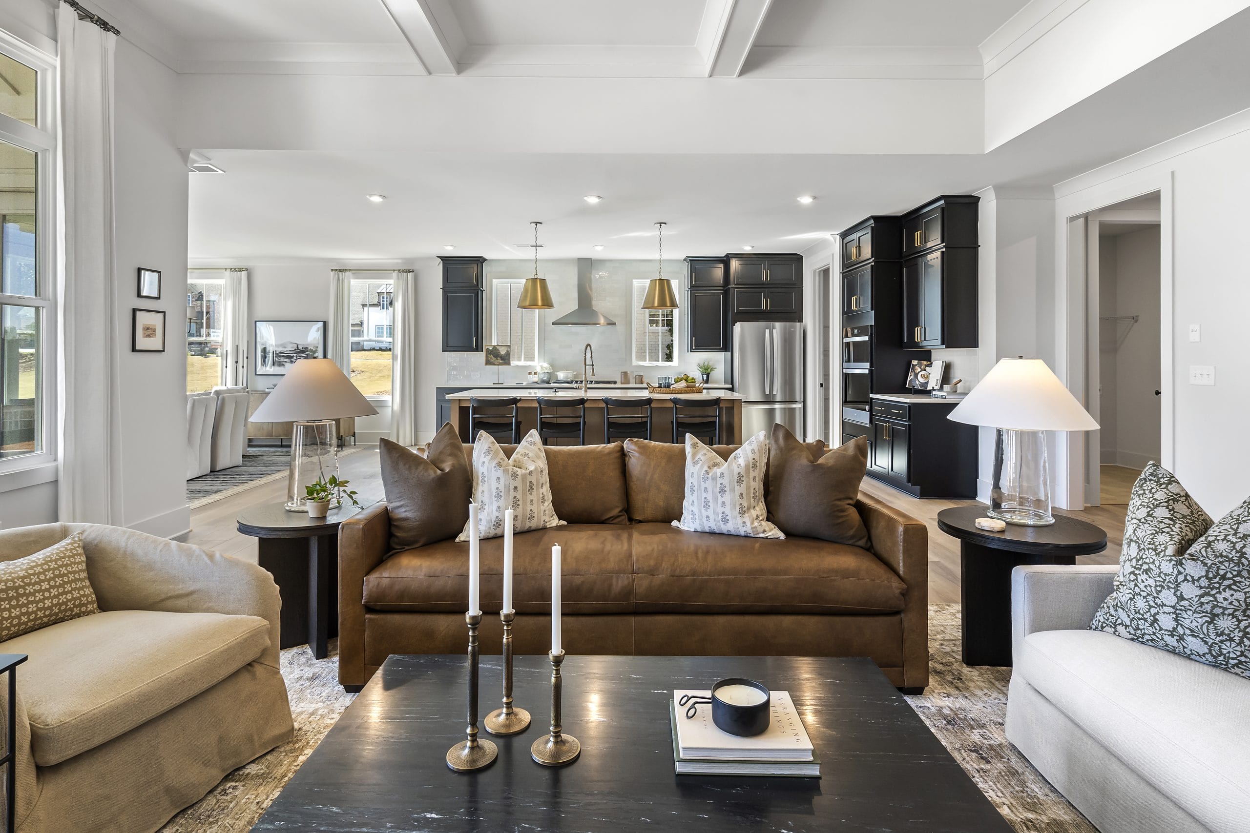 Modern open-concept living room and kitchen area with brown leather sofa, patterned throw pillows, round armchairs, black coffee table with decorative items, dark cabinetry, stainless steel appliances, pendant lighting, kitchen island with bar stools, and large windows.