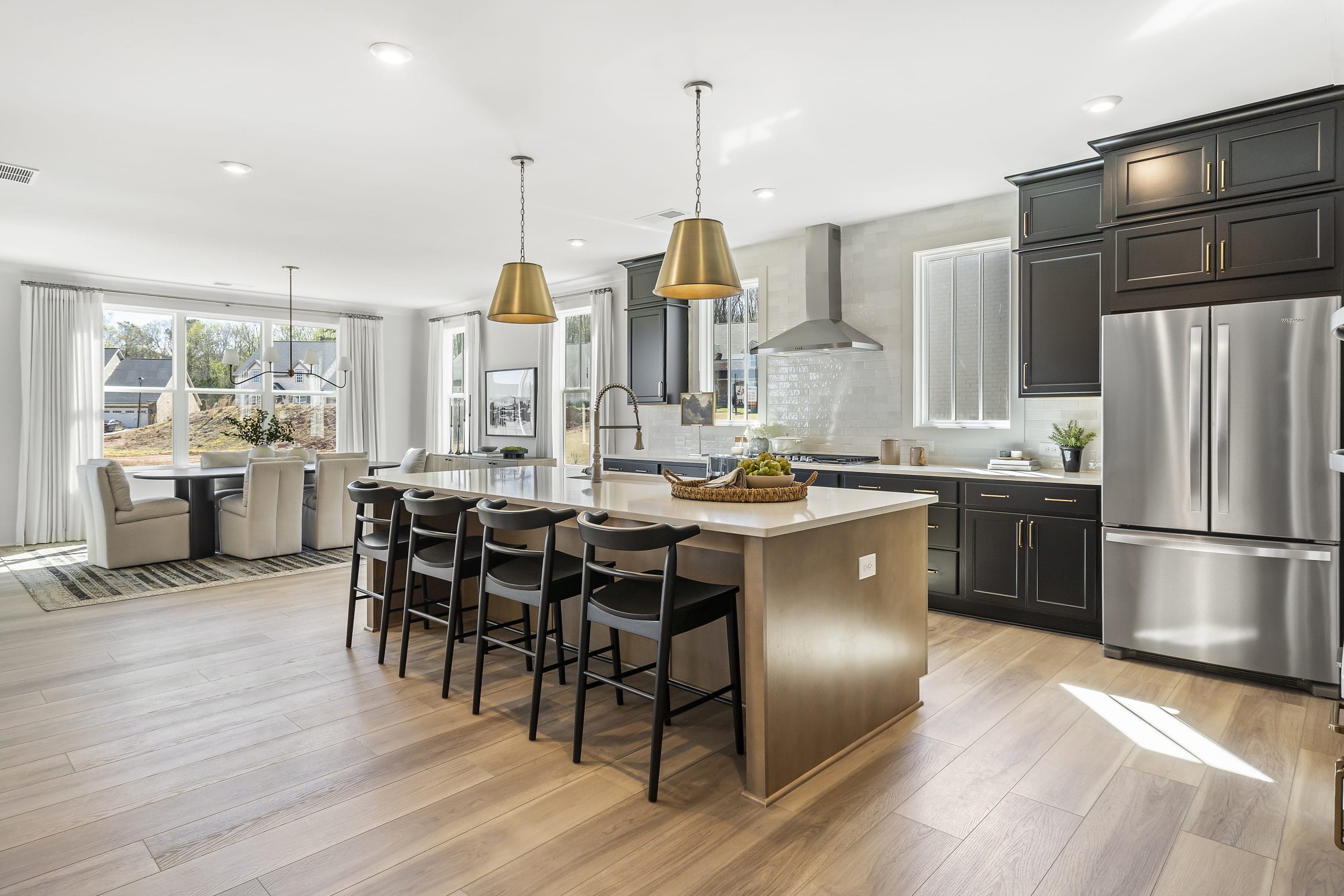 Modern kitchen and dining area in Heirloom , Cumming, GA, featuring large island, black chairs, pendant lights, stainless steel appliances, dark cabinetry, light wood flooring, dining table with six chairs near large windows.