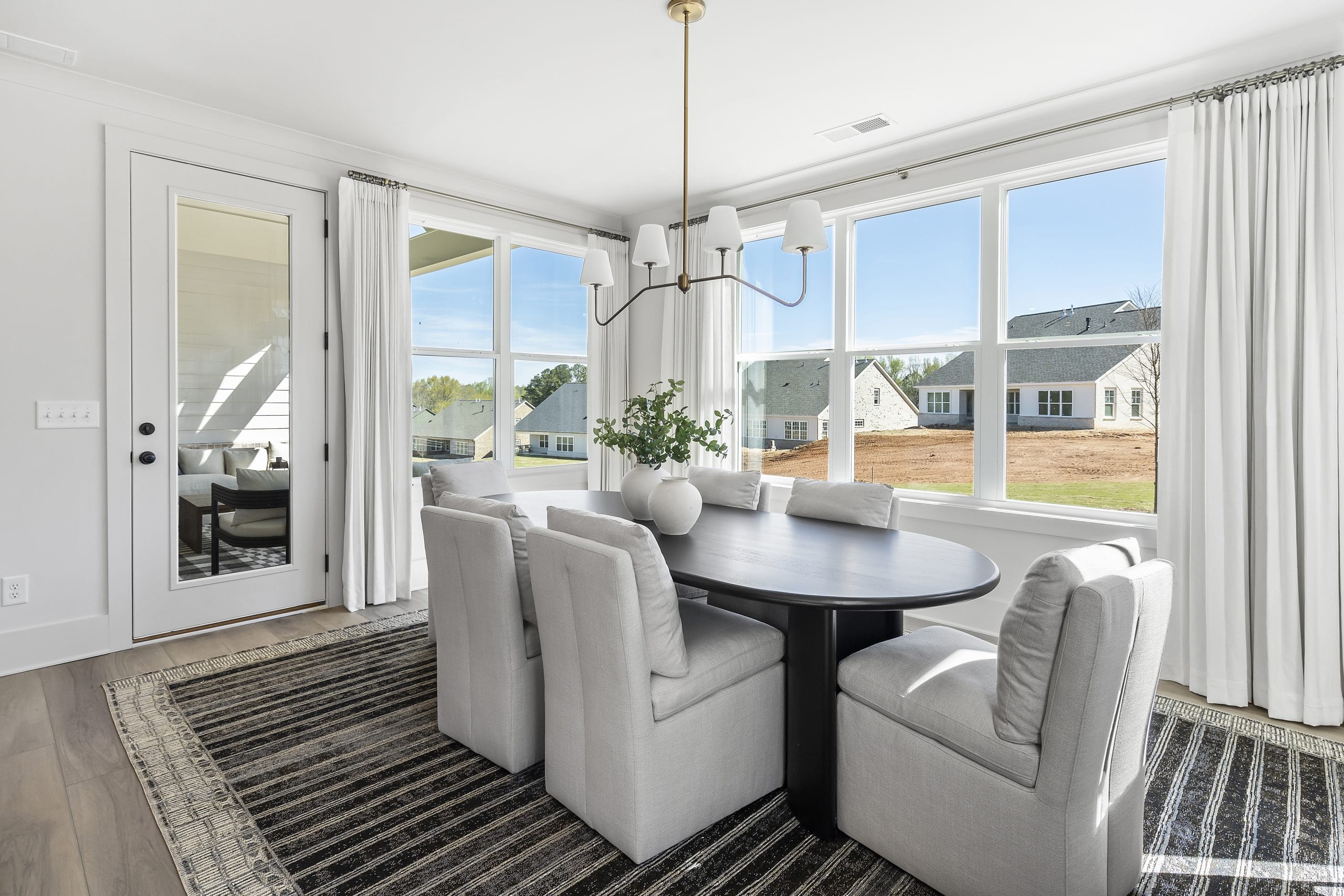 Modern dining room with large window, dark oval dining table, upholstered chairs, contemporary chandelier, white vase centerpiece, striped rug, white curtains, glass door.