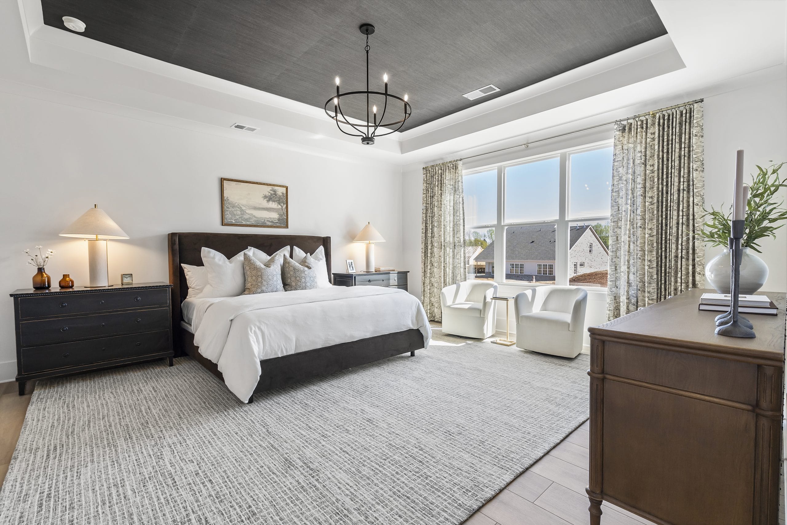 Modern bedroom featuring large bed with white bedding, dark nightstands with lamps, large window with patterned curtains, white chairs, small table, recessed ceiling with chandelier, dresser with decorative items, and area rug.