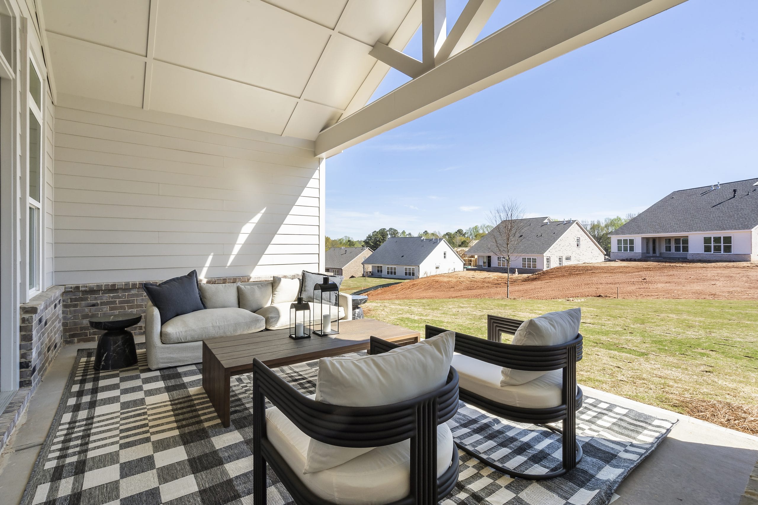 Covered outdoor patio with cushioned chairs, sofa, checkered rug, wooden coffee table, exposed beam roof, grassy yard, neighboring houses.