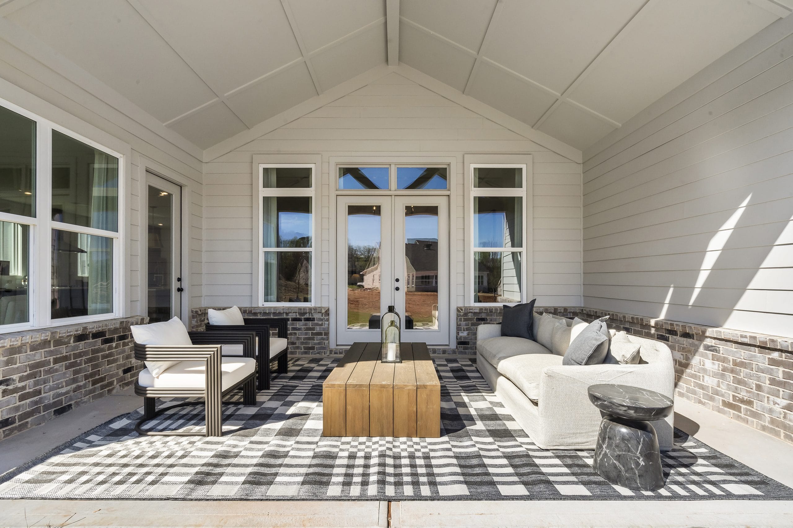 Covered outdoor patio with pitched roof, white sofa, armchairs, wooden coffee table, checkered rug, large windows, glass doors, light-colored walls with brick accents.