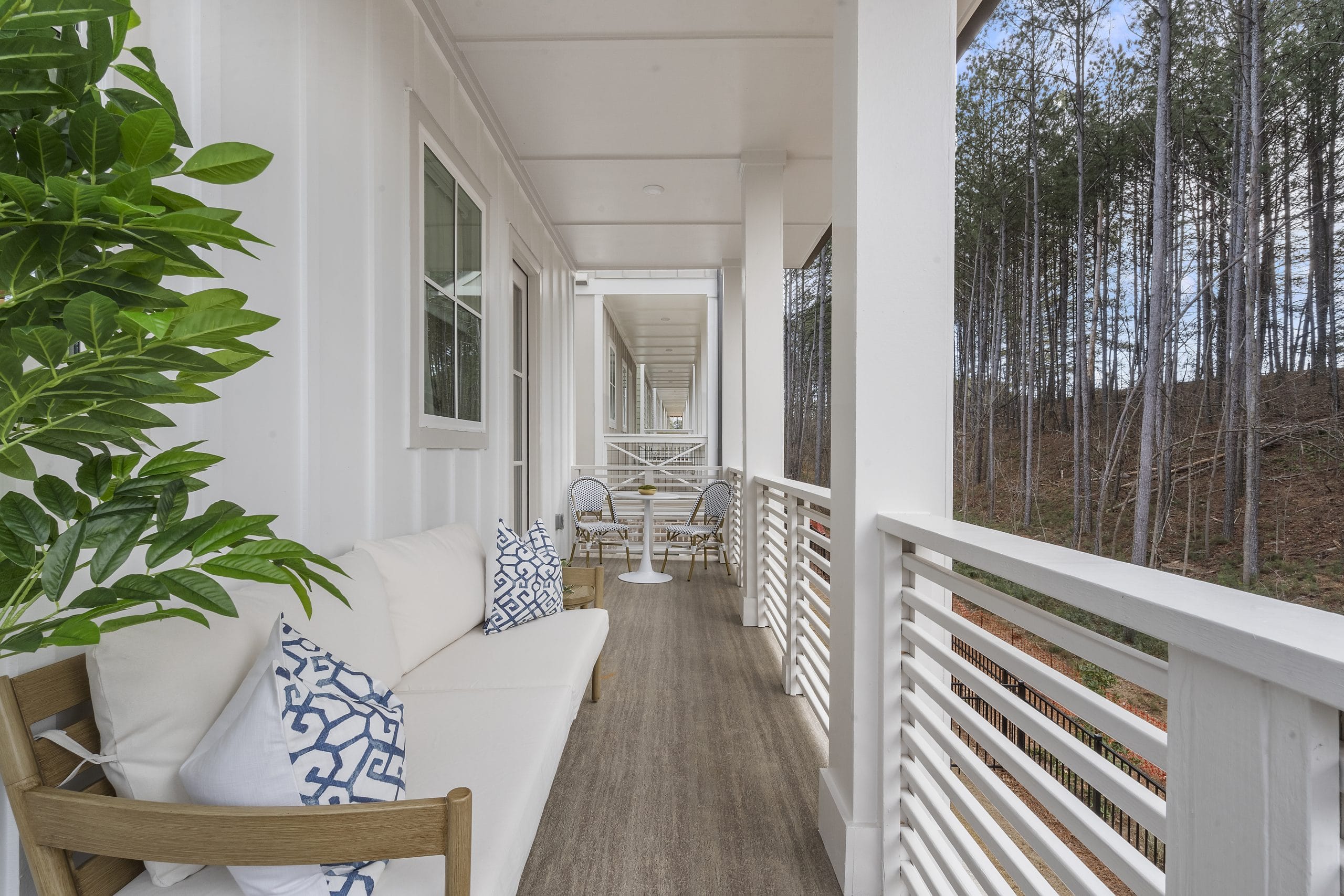 Outdoor patio with white furniture, a wooden bench with blue and white patterned cushions, potted plant, and a view of trees in the background.