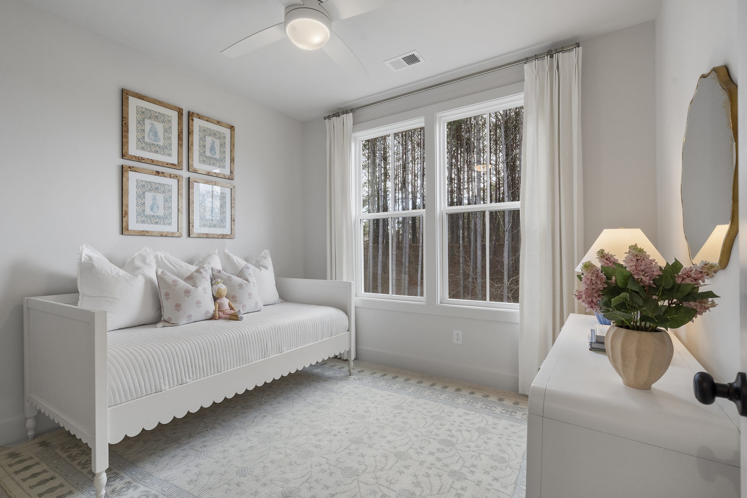Neatly arranged bedroom with a daybed against the left wall, adorned with white pillows and a stuffed animal, four framed pictures above the bed, large windows with sheer white curtains, white dresser with a round mirror and lamp with flowers, light-colored walls and carpet.