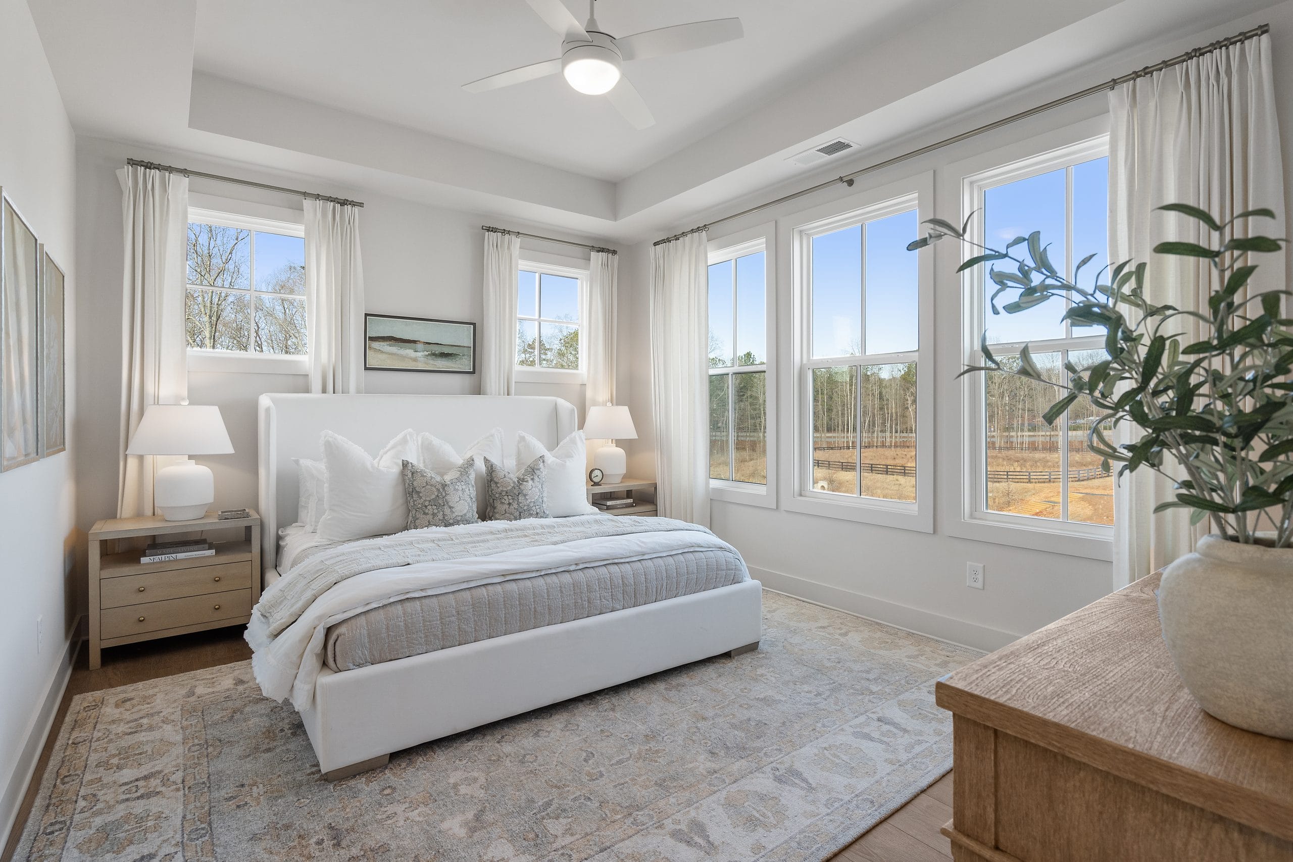 Well-lit bedroom with large windows, white bed with pillows and blanket, two bedside tables with lamps, dresser with a plant, white walls, and area rug on the floor.