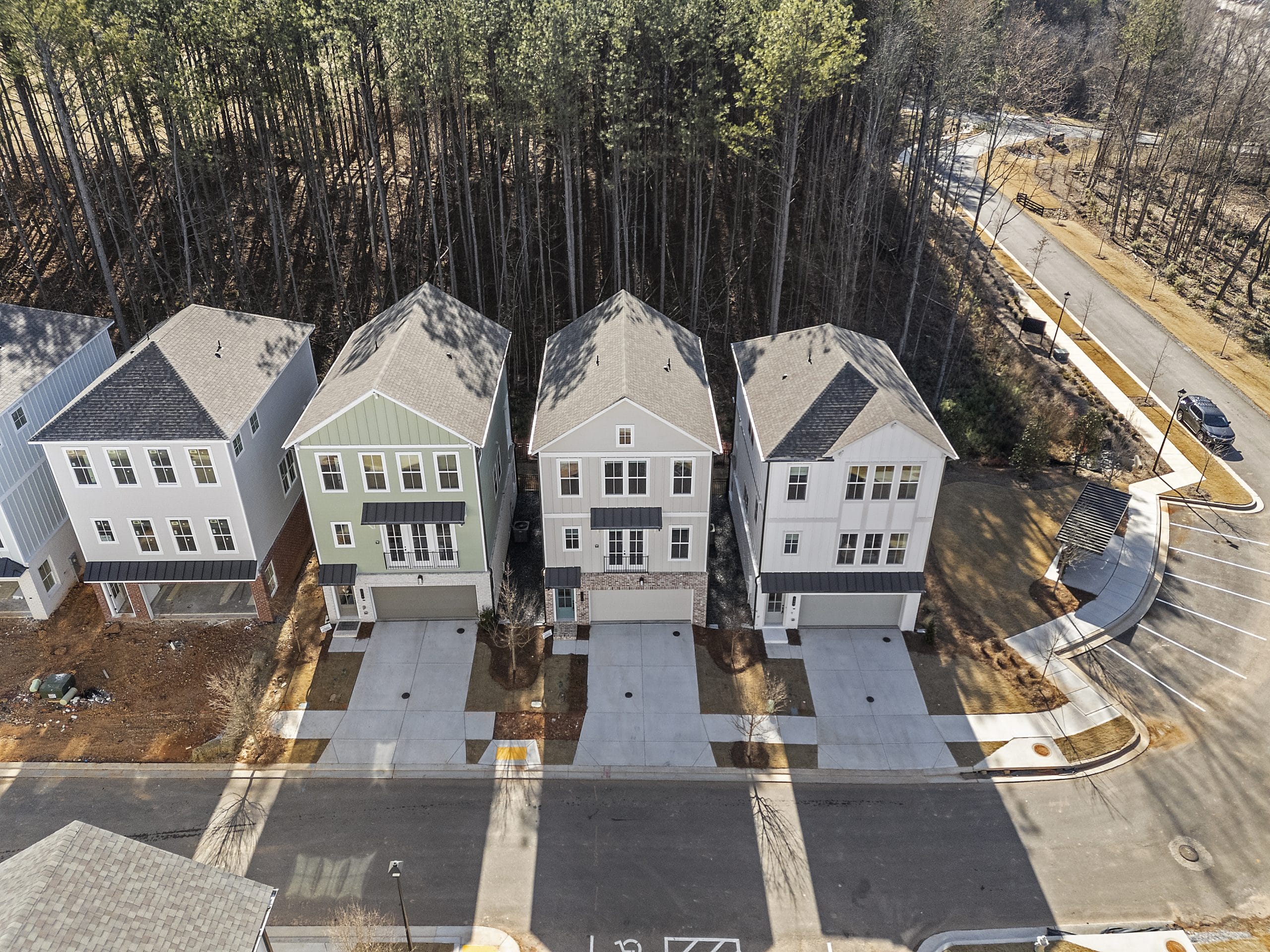 Aerial view of four modern two-story houses with garages, situated along a street in front of a wooded area.