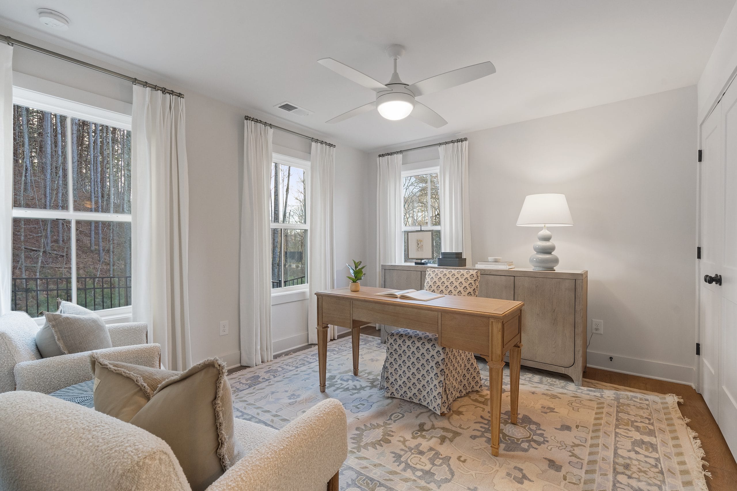 Room with large windows, white curtains, ceiling fan, wooden desk, two upholstered chairs, patterned rug, and sideboard with a lamp.