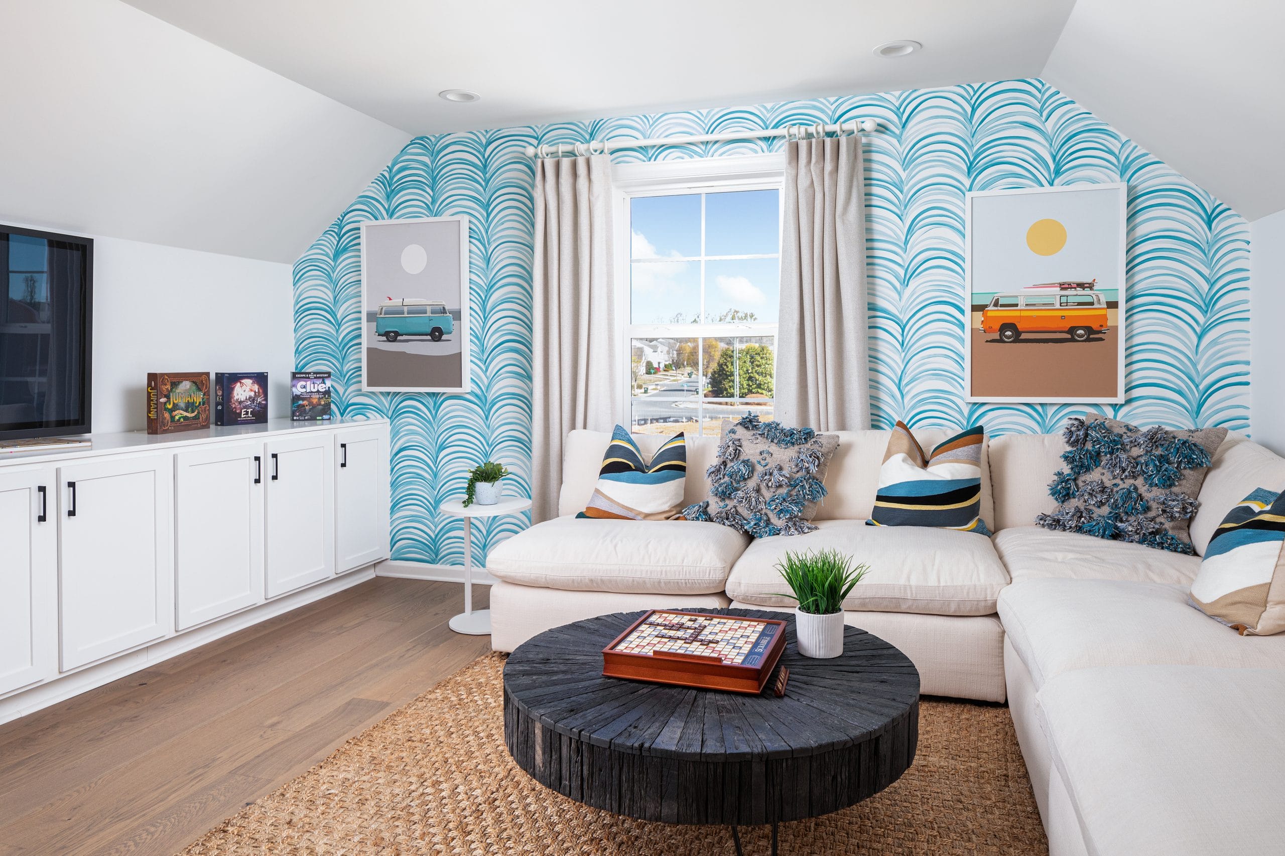 Bright and modern living room with white sectional sofa, patterned pillows, blue wave-patterned wallpaper, framed pictures, light-colored curtains, round black coffee table, built-in cabinet, flat-screen TV, wooden floor, and woven rug.