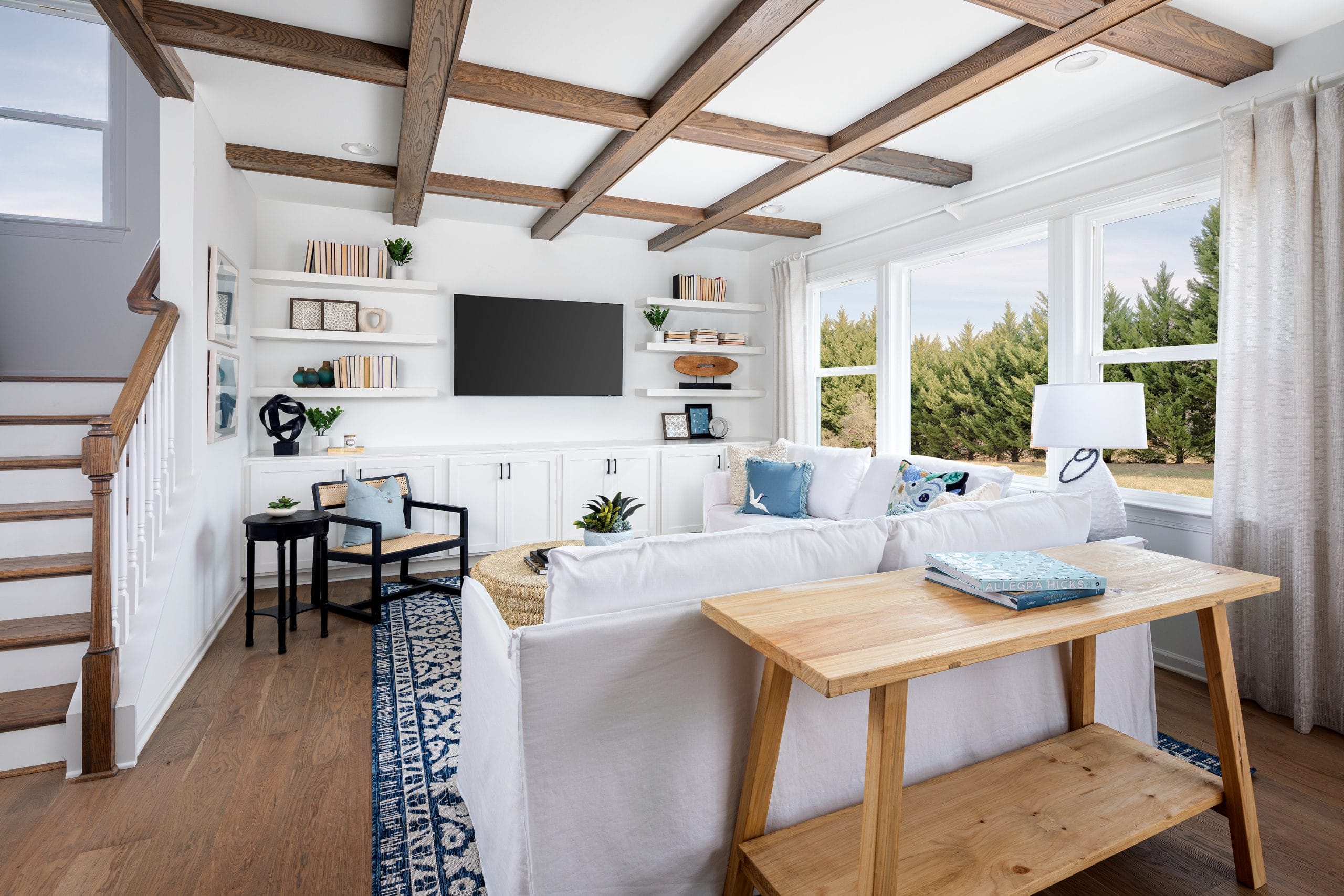 Family room with wooden ceiling beams, white walls, large windows, white sectional sofa with blue and white throw pillows, wall-mounted TV, built-in shelves, wooden coffee table, staircase, and blue patterned rug.