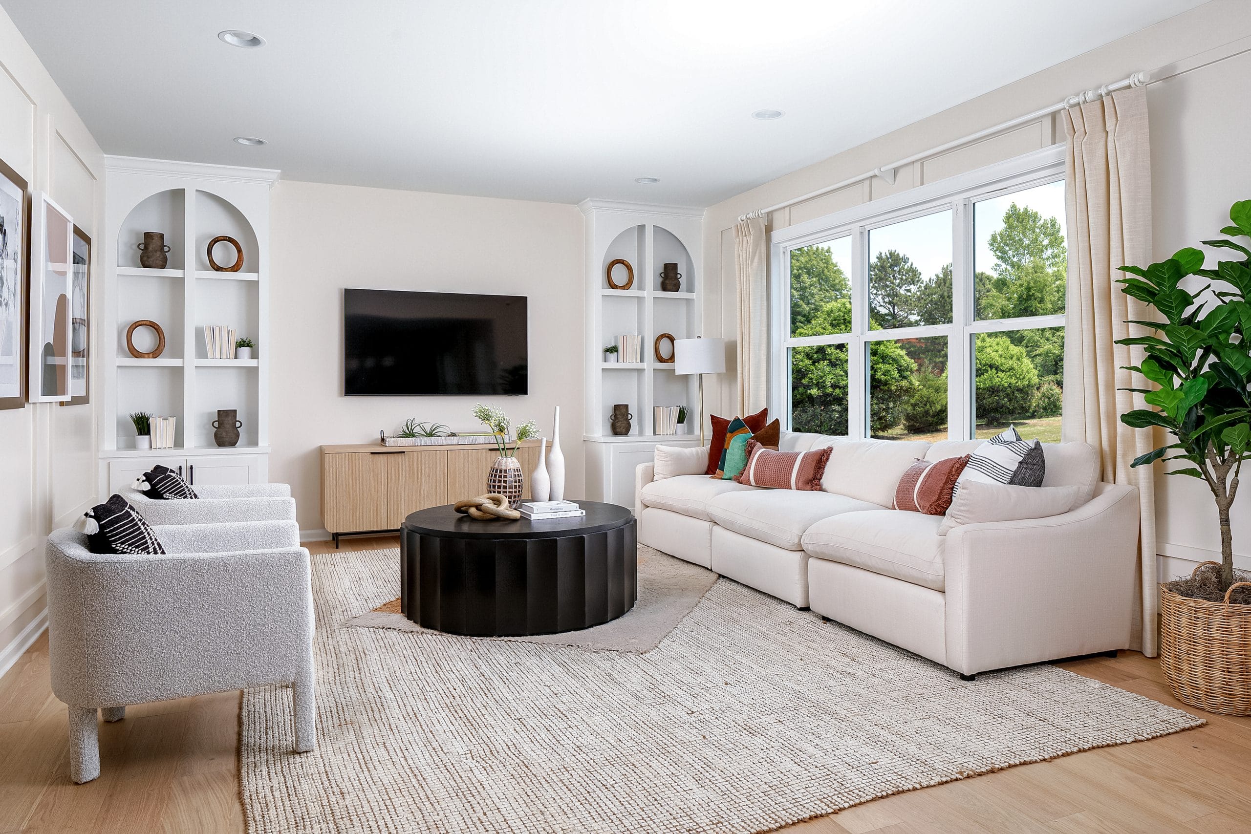 Modern living room with white sofa, colorful cushions, white armchairs, black round coffee table, wooden cabinet, wall-mounted TV, built-in shelves, and large green plant.