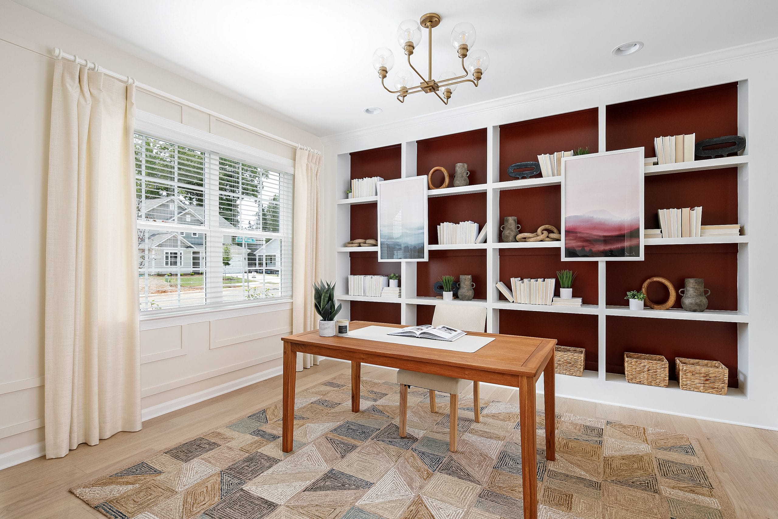 Well-lit home office with large window, white curtains, wooden desk, open book, small plant, built-in bookshelf, and patterned rug. The room is organized and inviting, perfect for productivity and focus.