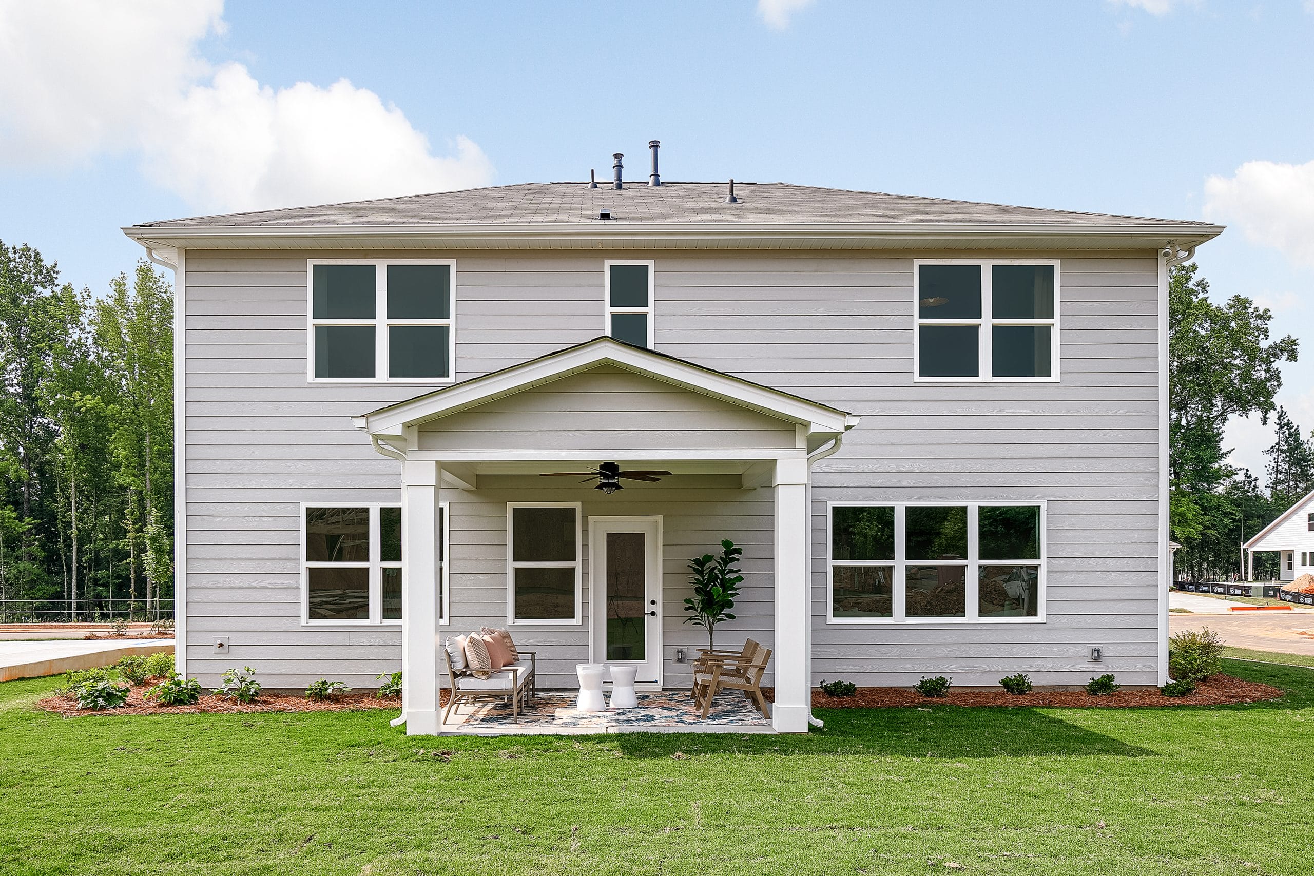 Back view of a two-story house with light gray exterior, multiple windows, covered patio with outdoor furniture, grassy lawn, landscaped garden beds, and trees in the background.