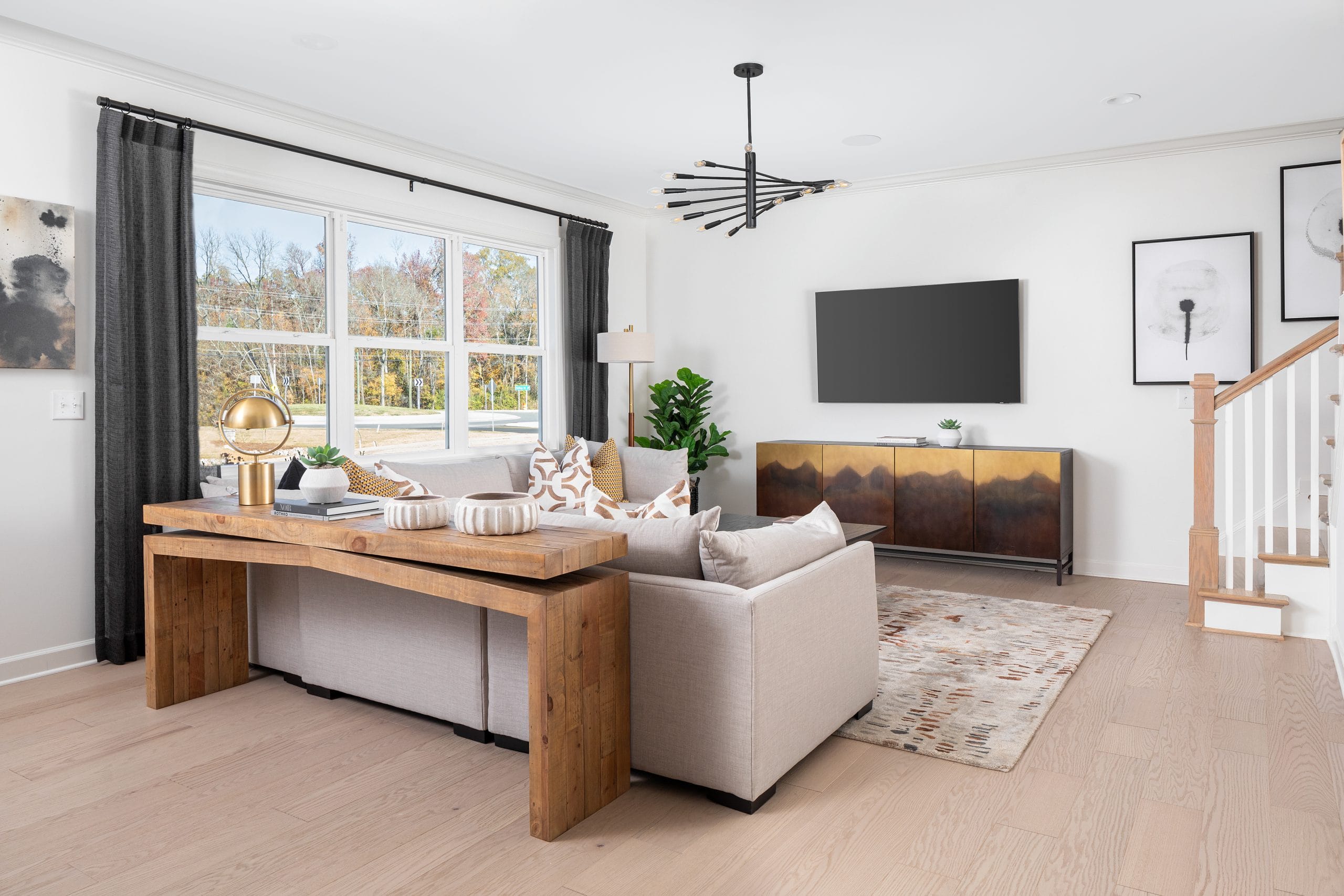 Modern living room with beige sectional sofa, patterned throw pillows, wooden console table, and large window with dark curtains. Black chandelier, staircase, cabinet with artistic design, flat-screen TV, and framed artworks.