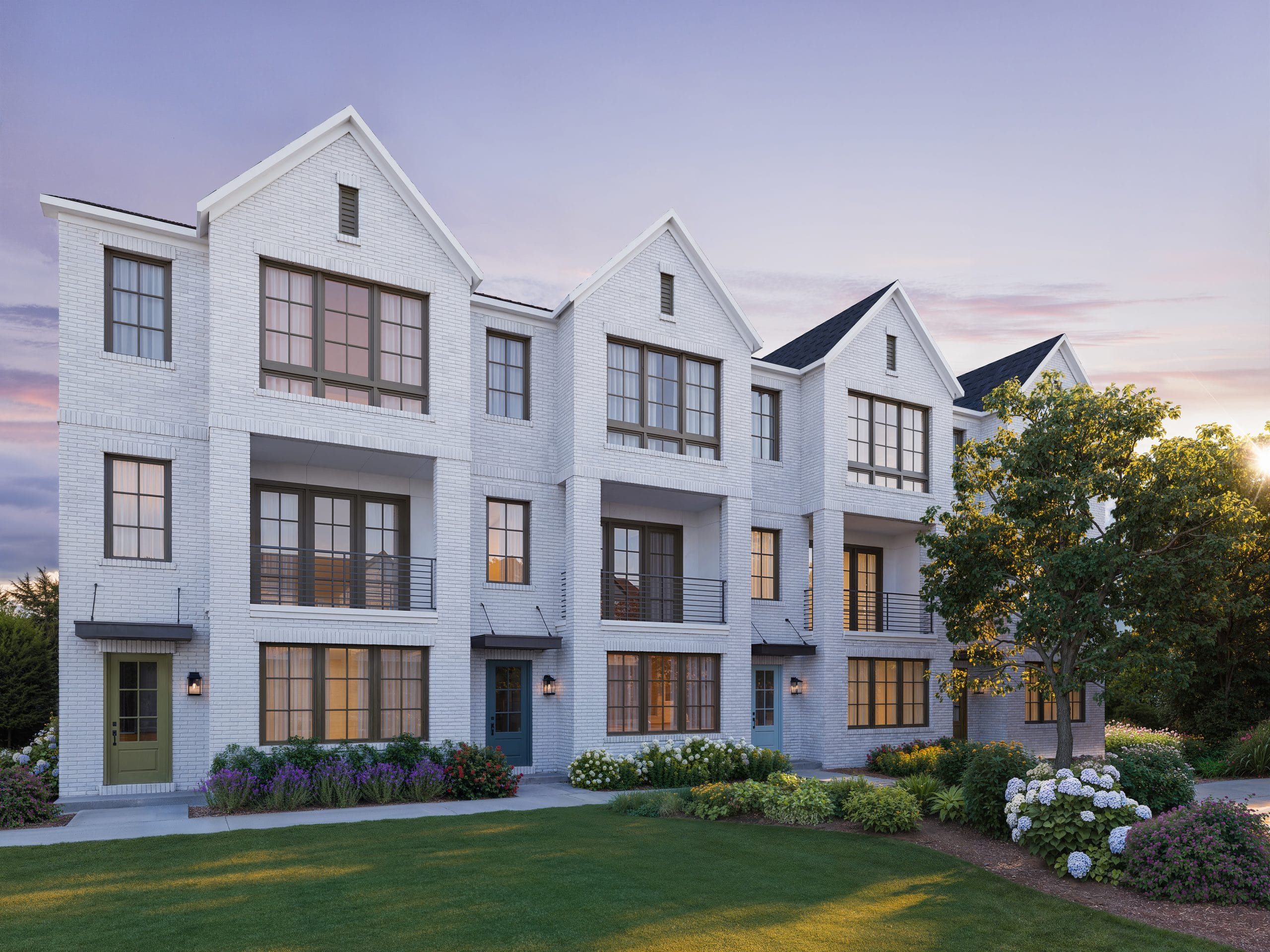 Modern multi-story residential building with white brick exterior, large windows, balconies, multiple entrances, landscaped front with grass, bushes, and flowering plants, clear sky with sunset colors.