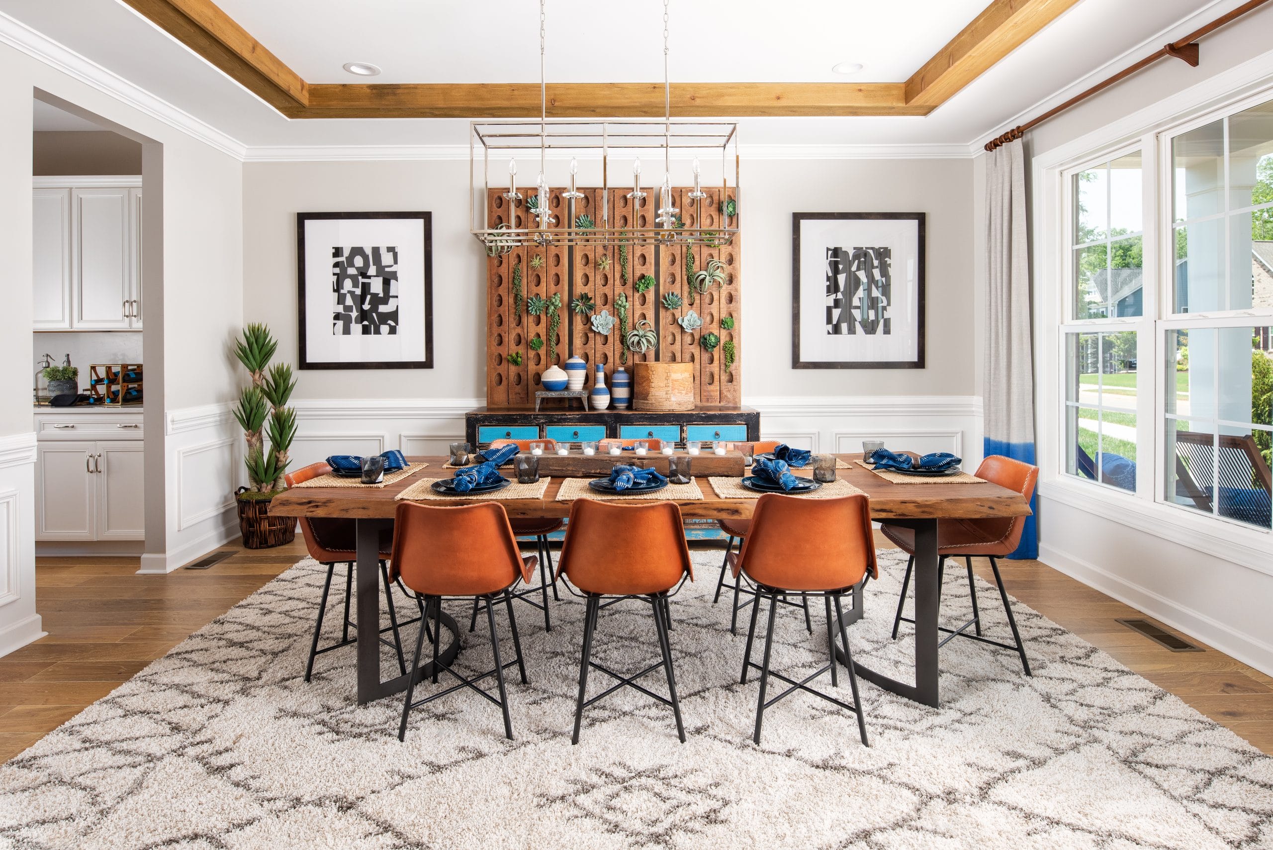 A dining room with a large wooden table surrounded by eight brown chairs. The table is set with blue napkins and plates. Two framed artworks and a decorative wooden panel with hanging plants adorn the wall behind the table. The room has white walls, a patterned rug, and large windows on the right side allowing natural light to enter. A small plant is placed in the corner near the left side of the image.