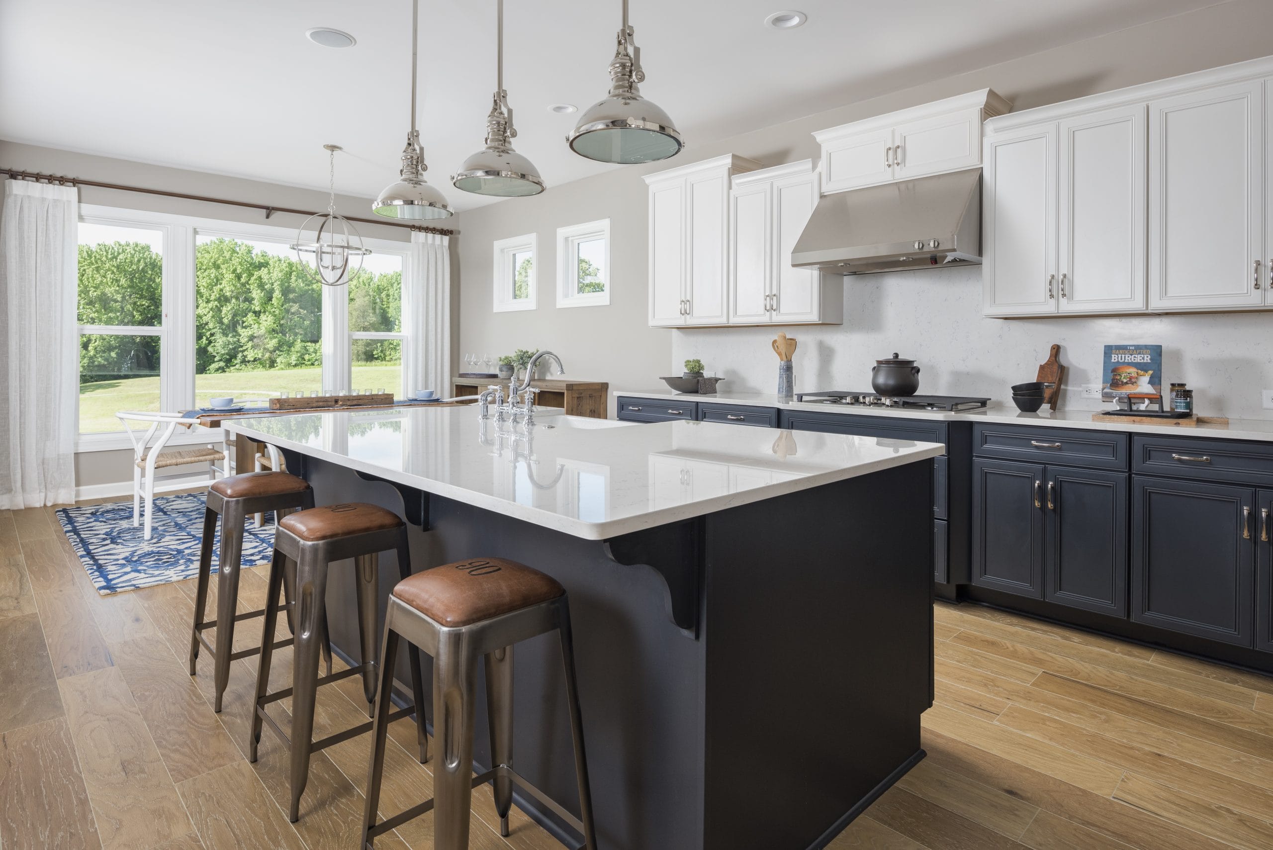A modern kitchen with a large island in the center. The island has a white countertop and dark blue cabinetry, with three brown leather bar stools placed along one side. Above the island, there are three pendant lights with metallic shades. The kitchen features white upper cabinets and dark blue lower cabinets, along with stainless steel appliances including a range hood and stove. There is a window above the sink area and another set of windows providing natural light from the left side of the image. The floor is made of light-colored wood, and there is a dining area visible in the background with a table and chairs on a blue patterned rug.