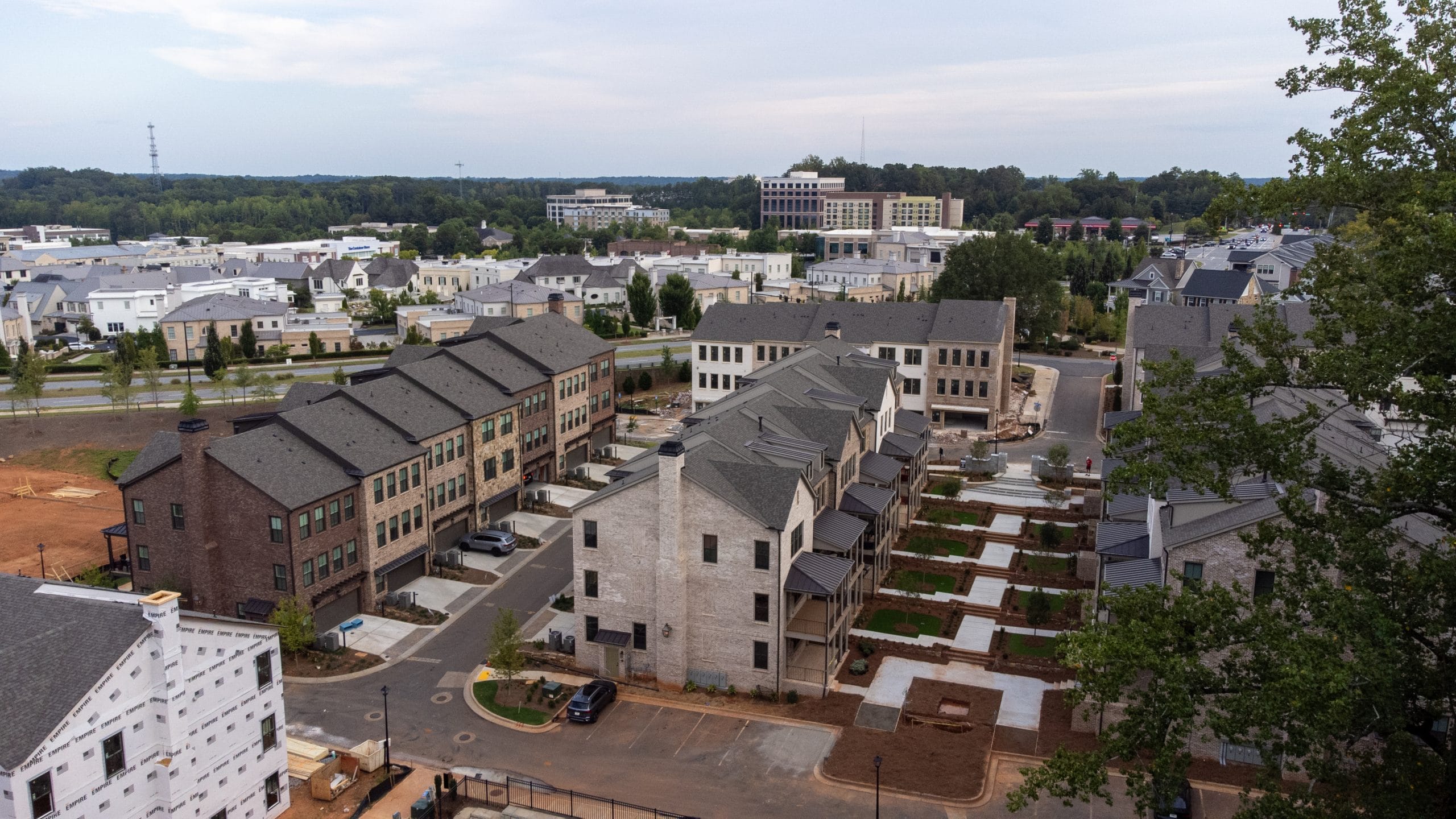 Residential area with multiple rows of townhouses and apartment buildings, orderly arrangement with streets and sidewalks, ongoing construction with materials and unfinished structures, background with more residential and commercial buildings, surrounded by greenery including trees and grass patches.