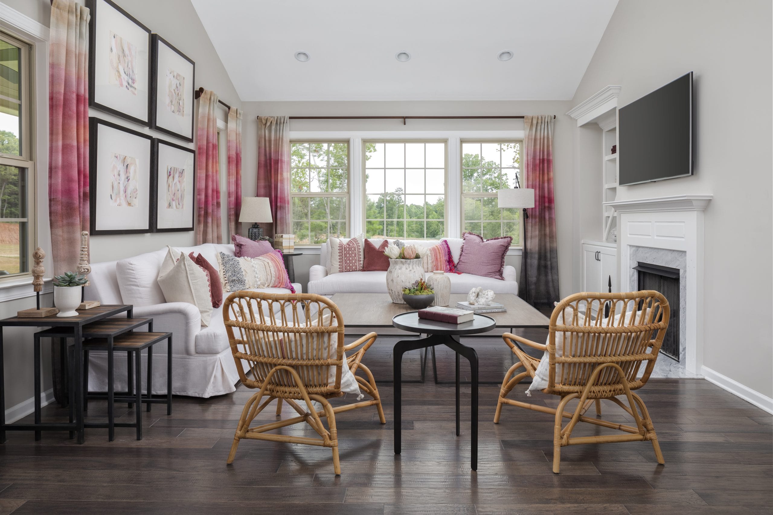 Modern living room with white sofa, pink and white cushions, wicker chairs, round black coffee table, side tables, framed artwork, flat-screen TV above white fireplace, dark wood floor, and pink and beige curtains.